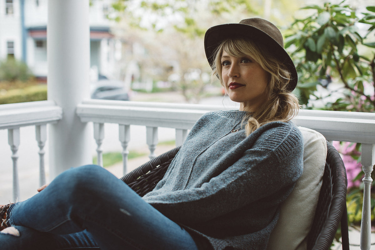 A woman sitting on the front porch of a house, modeling the sweater called Weekender Light